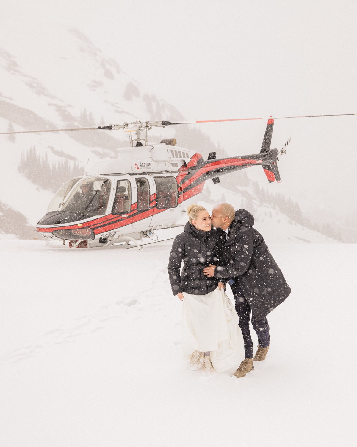 A couple shares a tender moment in the snow, with a helicopter parked nearby, surrounded by a winter landscape.