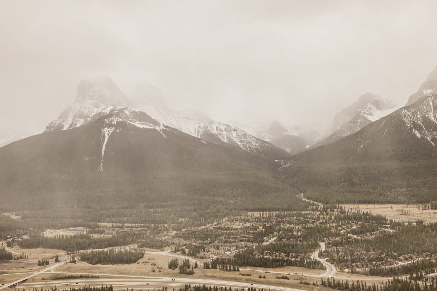 A serene view of mountains with snow-capped peaks, surrounded by dense forests, under a hazy sky.