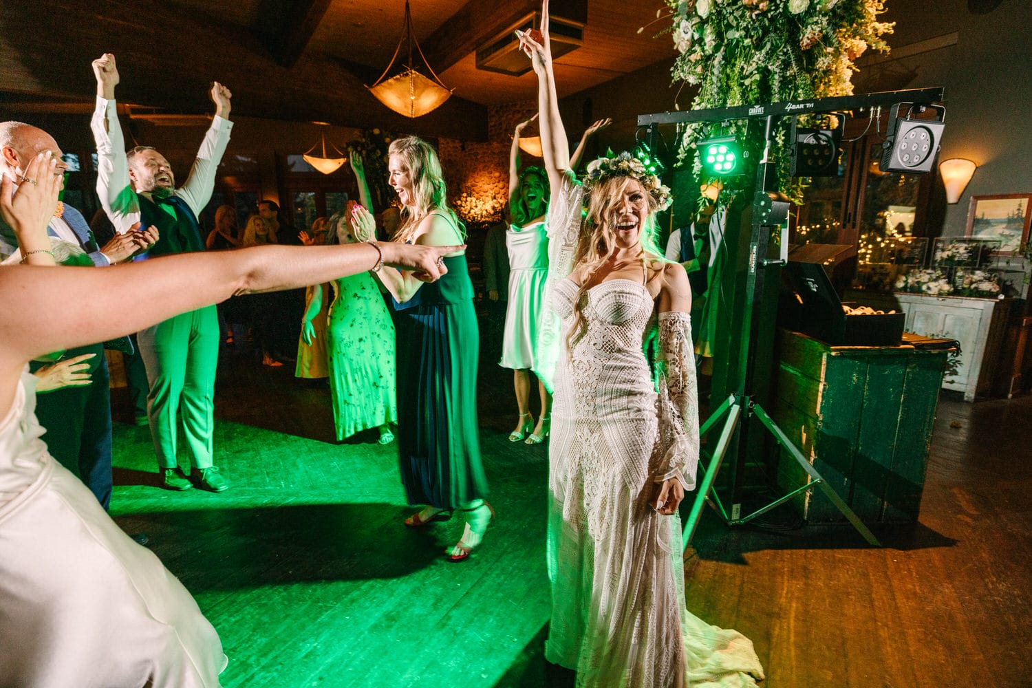 A joyful bride in a stunning white gown leads the dance floor with raised arms, surrounded by guests enjoying the festive atmosphere with green lighting.