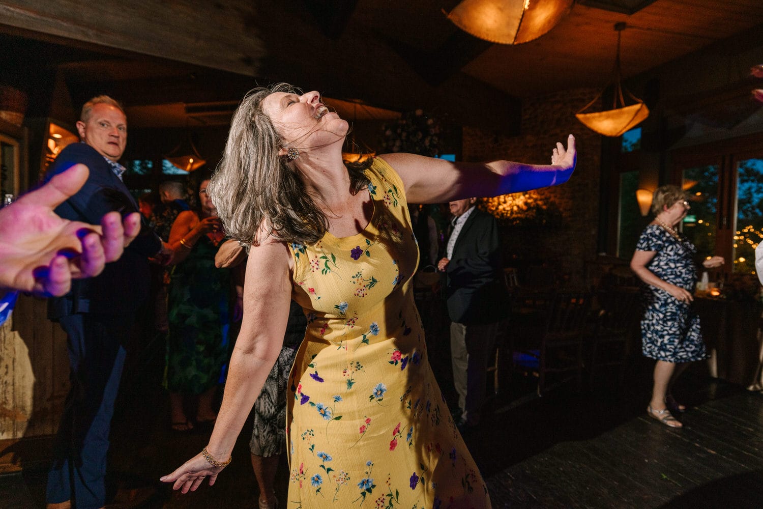 A woman in a floral yellow dress dances energetically, surrounded by guests enjoying a festive atmosphere.