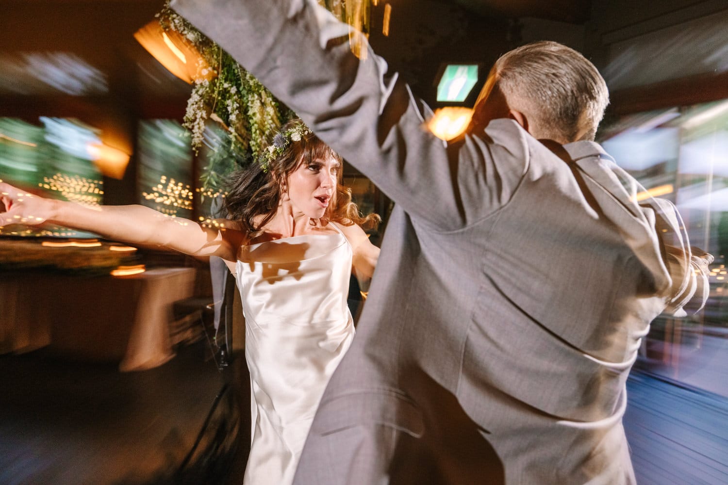 A joyful bride twirls energetically with a guest, surrounded by a blurred backdrop and twinkling lights, capturing the festive atmosphere of the celebration.