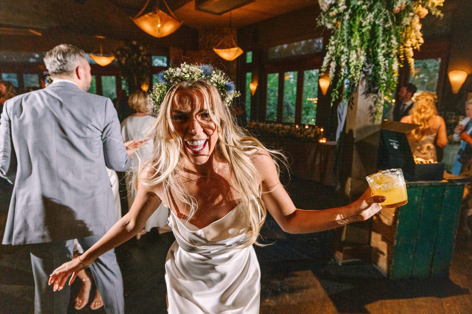 A bride in a flowing white dress and floral crown smiles widely while holding a drink, surrounded by guests celebrating at a lively party.