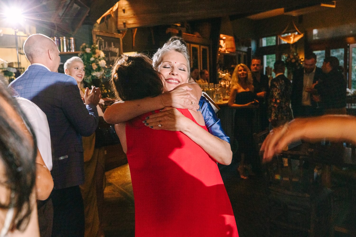 Two women joyfully hugging at a lively event, surrounded by friends and a warm atmosphere.