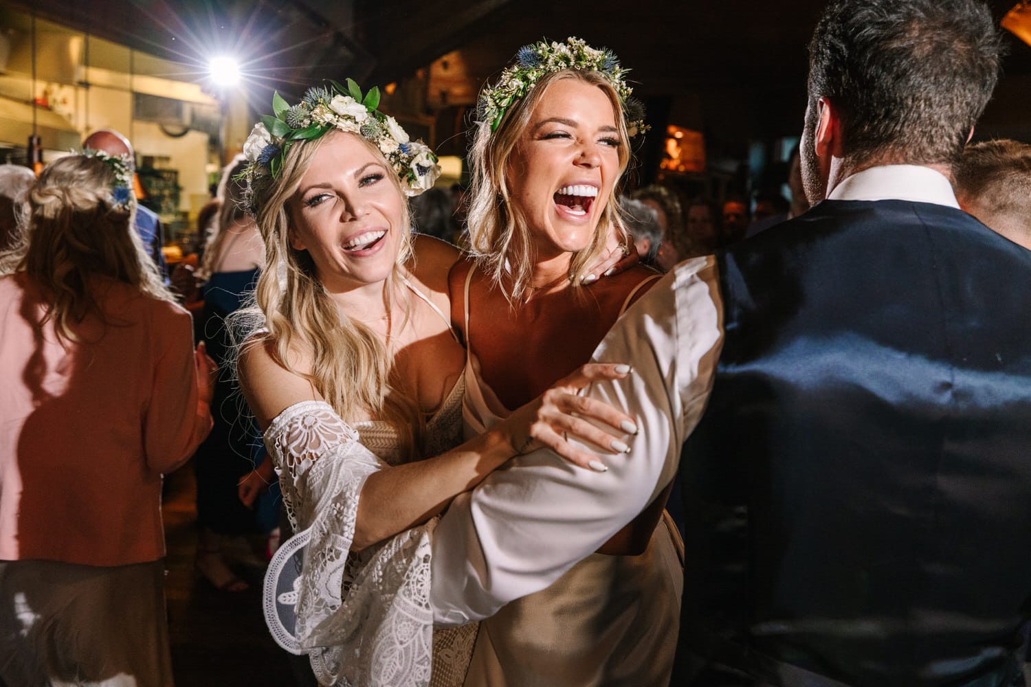 Two women in floral crowns enjoy a lively moment on the dance floor, surrounded by guests and festive atmosphere.