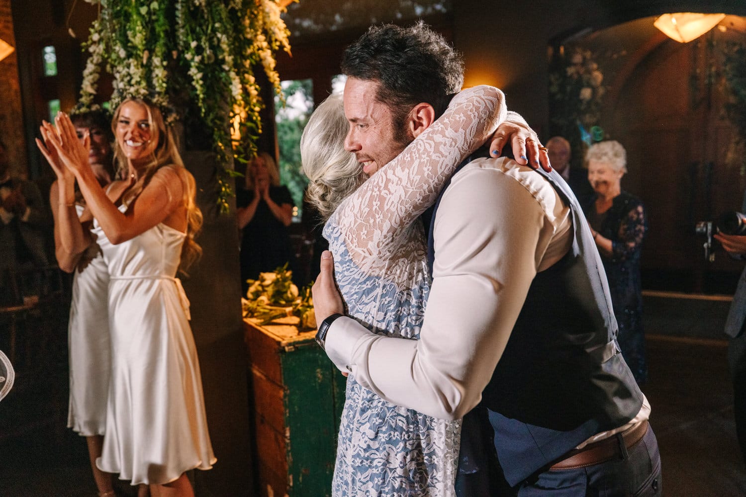 A joyful moment showcasing a man embracing a woman in a floral-filled venue, with guests clapping and smiling in the background.