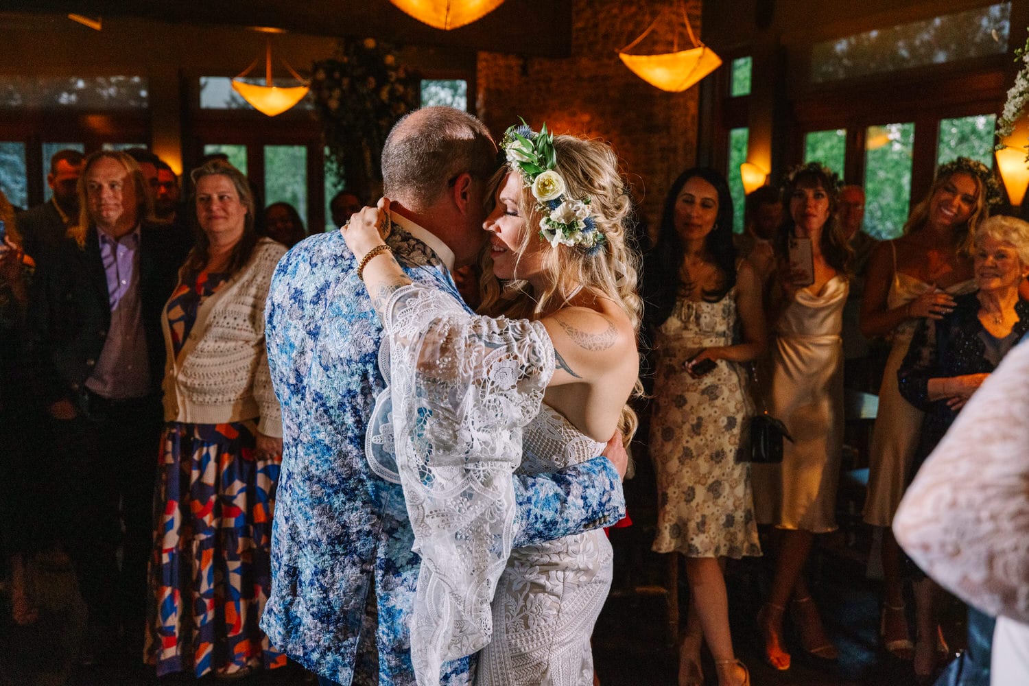 A couple shares an intimate dance surrounded by friends and family at a wedding reception. The bride, wearing a white gown adorned with floral accents, embraces the groom, who is dressed in a colorful floral jacket, while guests watch and celebrate.