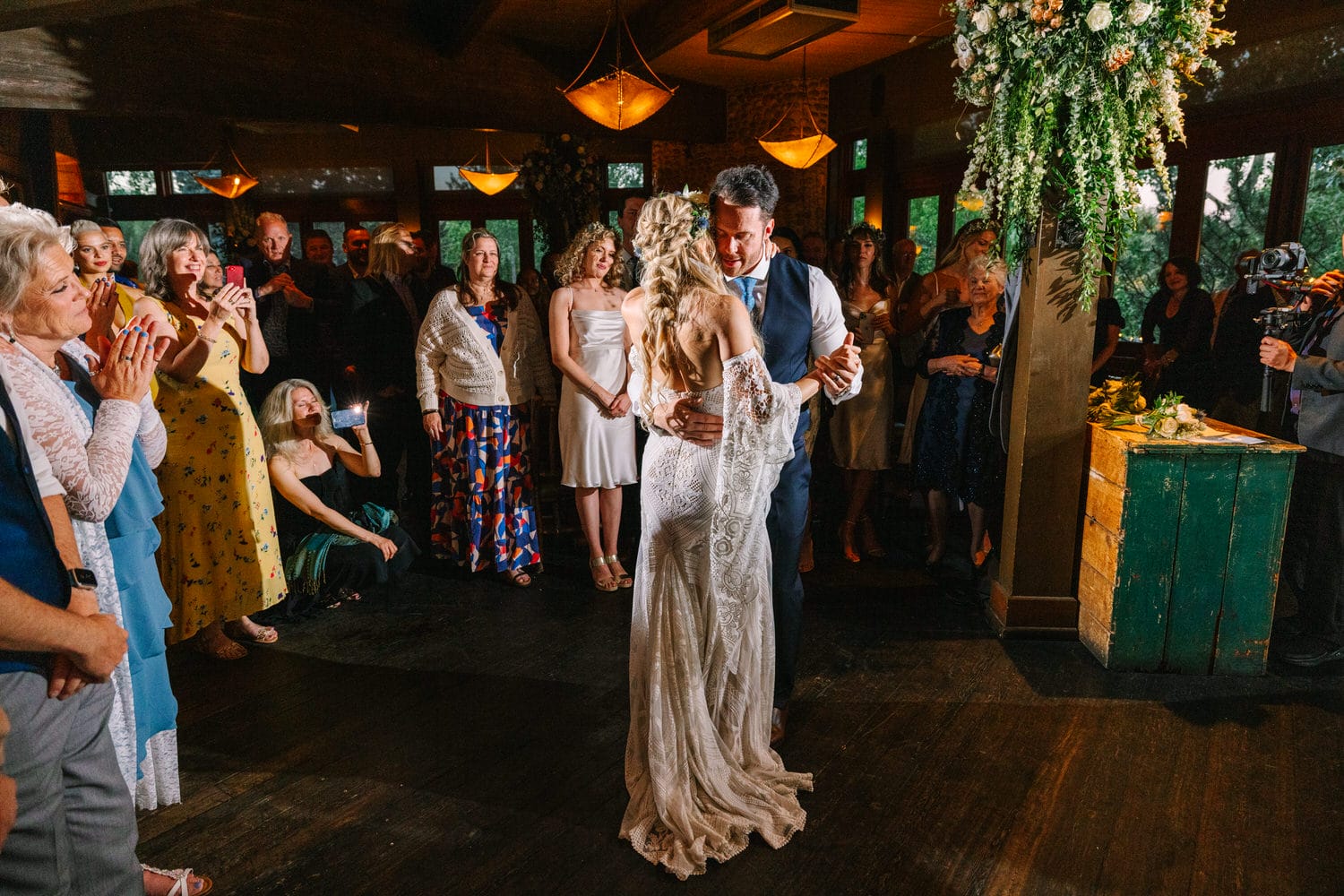 A bride and groom share an intimate dance, surrounded by joyful guests capturing the moment on their phones, in a warmly lit venue decorated with floral arrangements.