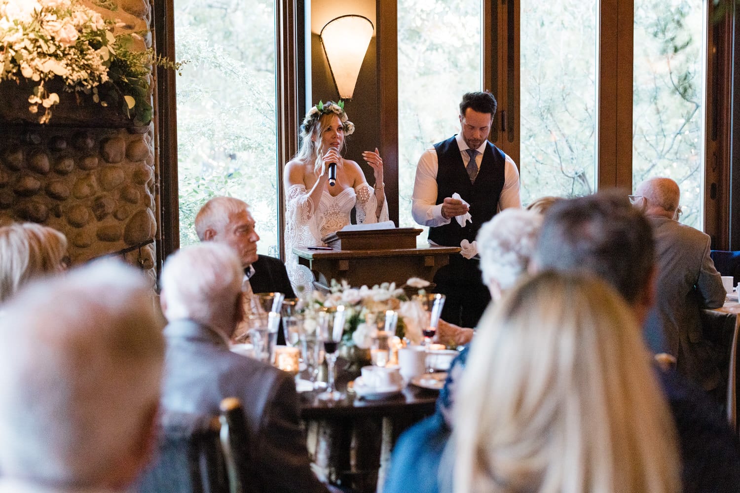 A woman in a flower crown speaks at a wedding reception while a man stands beside her, preparing to assist. Guests seated at a round table listen attentively.