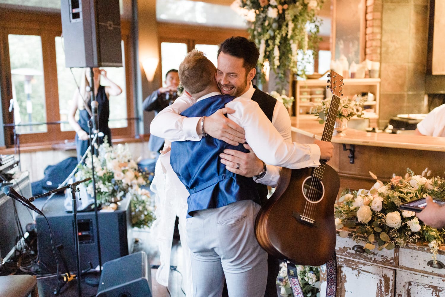 Two men share a warm hug in a beautifully decorated venue, with one holding an acoustic guitar, surrounded by floral arrangements.