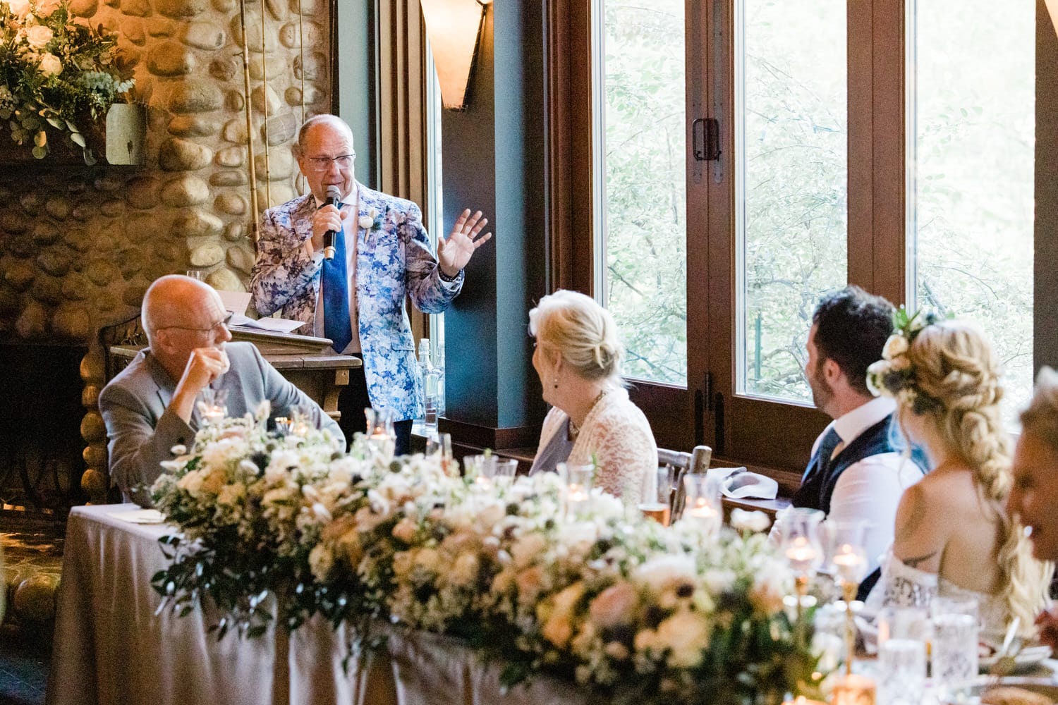 A speaker in a floral jacket shares a heartfelt toast at a wedding reception, while the seated guests enjoy the moment amidst elegant floral arrangements.