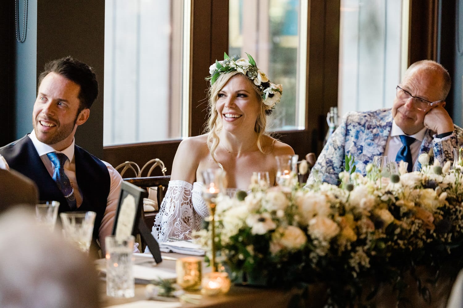 A bride in an off-shoulder gown with a floral crown smiles as she enjoys the company of guests at a festive wedding reception.