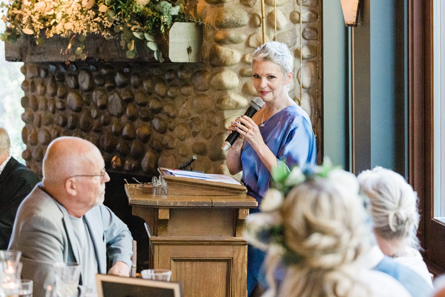 A woman in a blue dress speaks into a microphone at a gathering, with guests attentively listening in a warmly decorated venue.