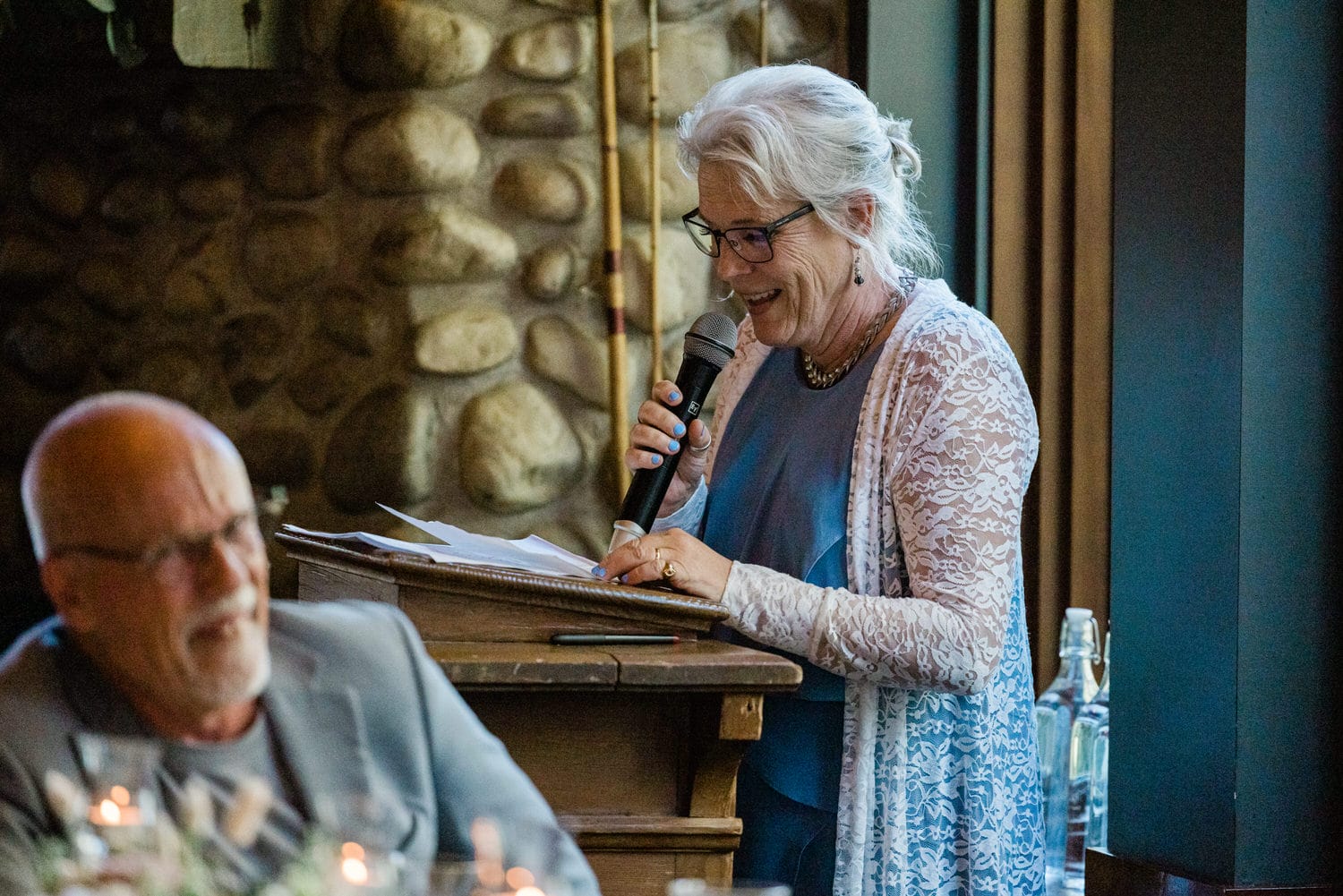 A woman in glasses smiles as she speaks into a microphone, while a man in the foreground listens, against a backdrop of a stone wall.