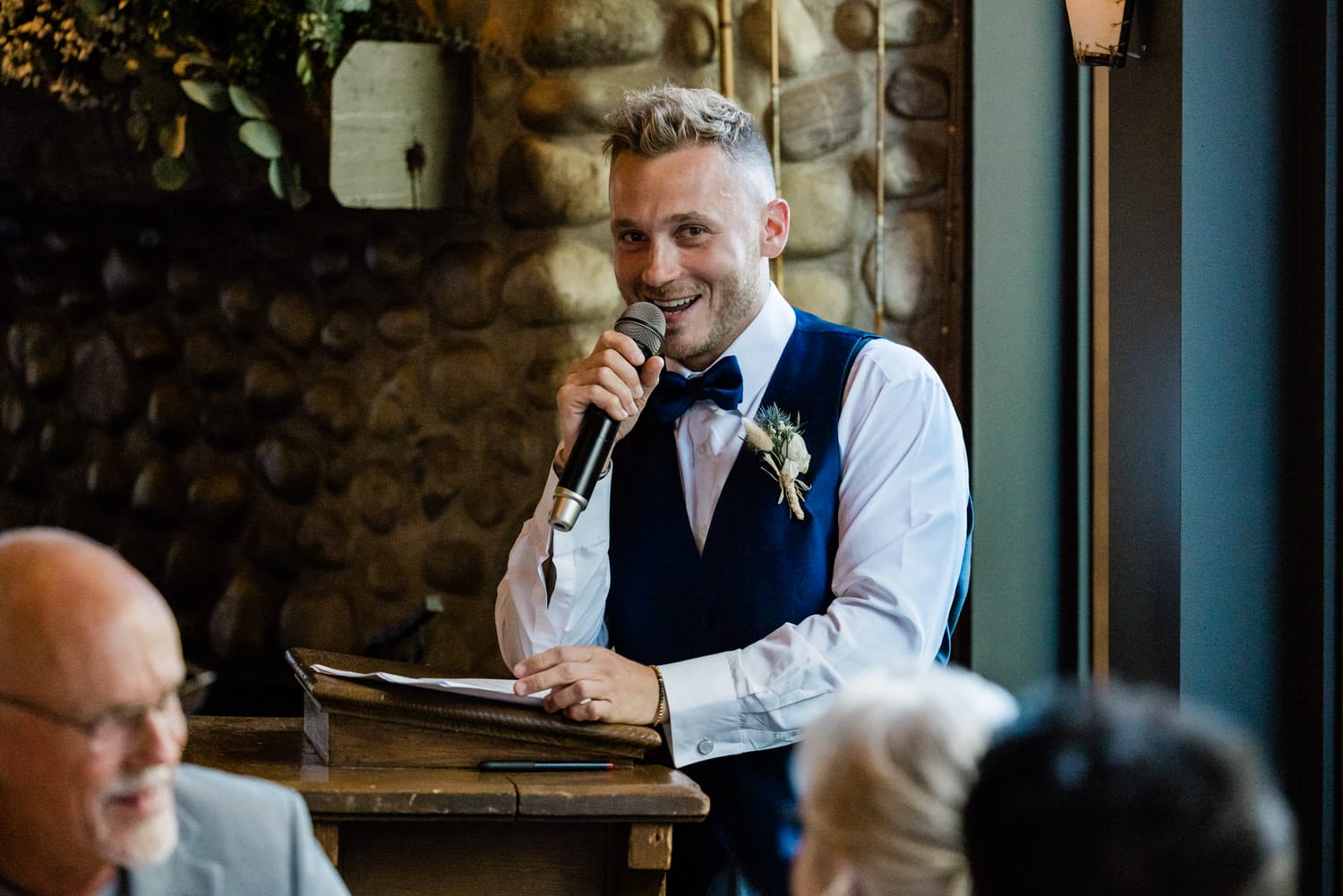 Man in a blue vest and bow tie speaking into a microphone during a wedding reception, with guests seated around him.