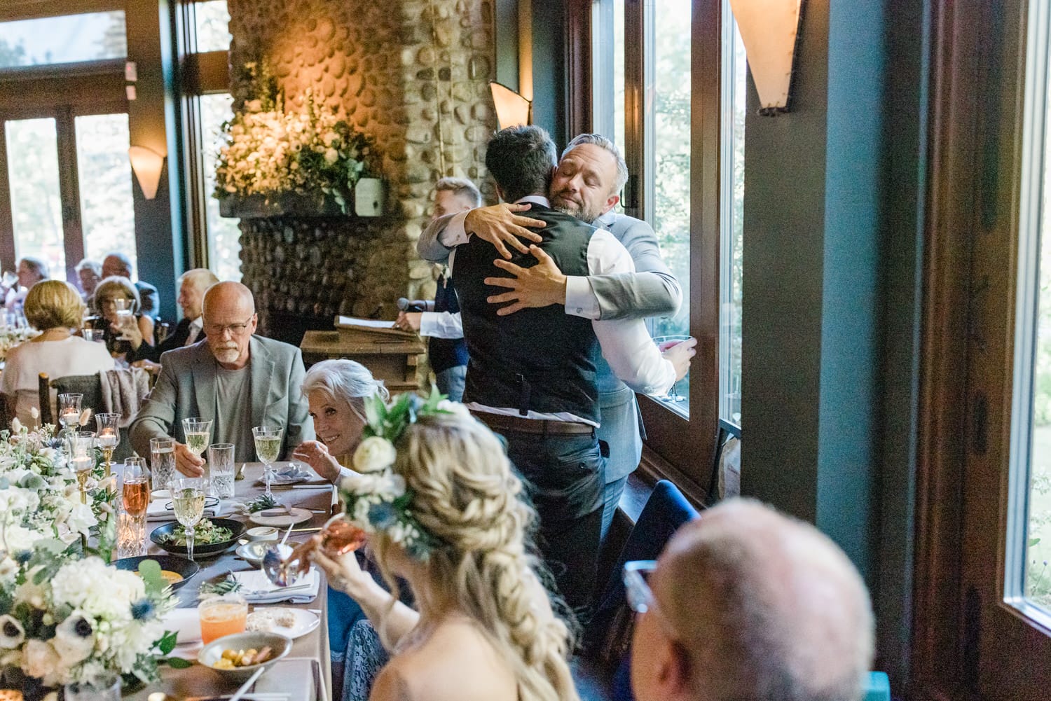 A joyful moment of two men embracing during a wedding reception, surrounded by guests seated at a beautifully decorated table with floral arrangements and drinks.