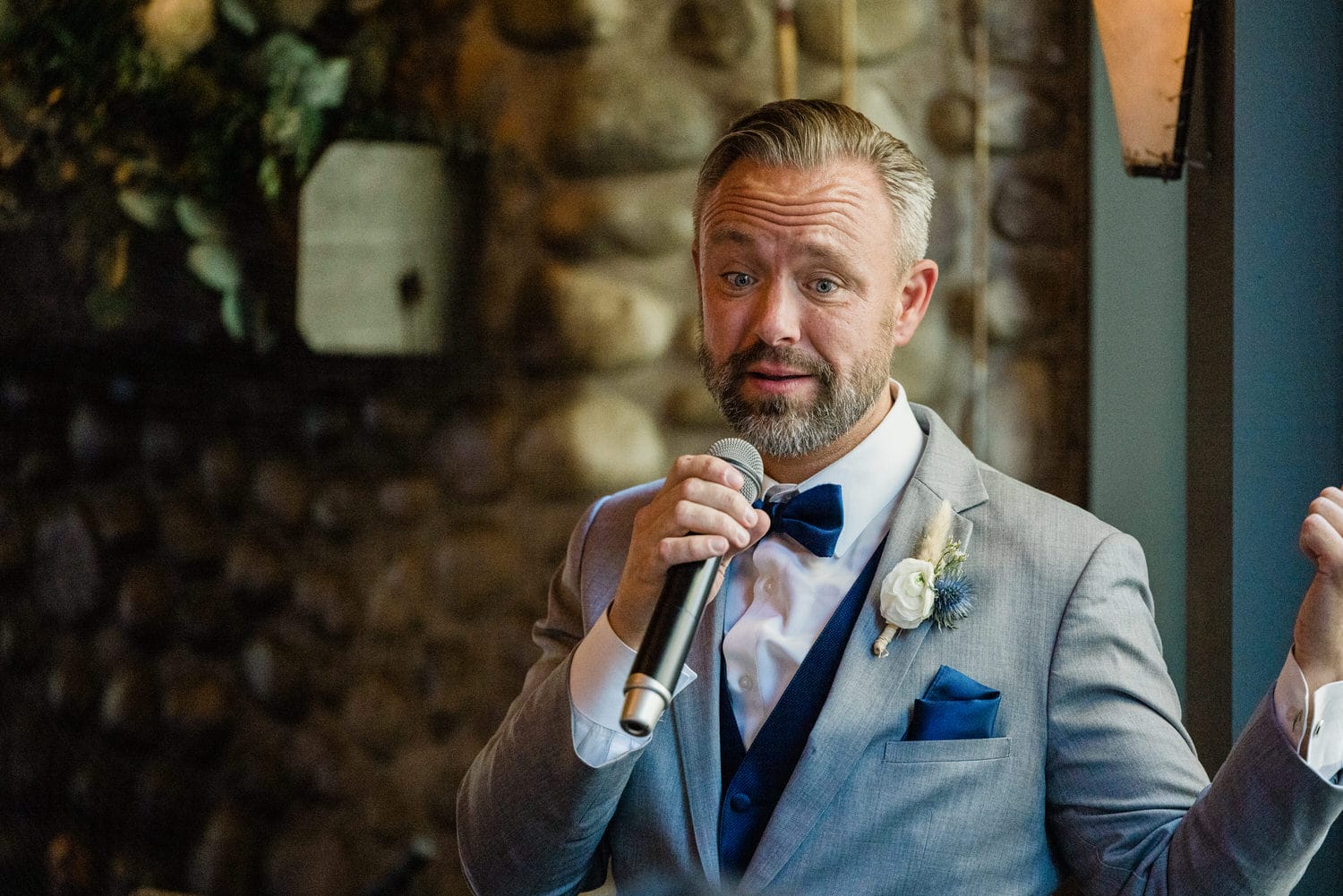 A man in a gray suit and blue bow tie passionately delivers a speech while holding a microphone, set against a rustic stone backdrop.