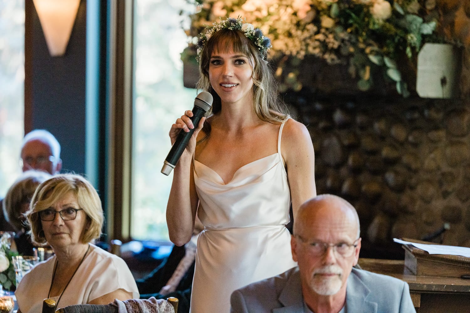 A woman in a white dress with a floral headband delivers a speech while holding a microphone, with guests attentively listening in the background.