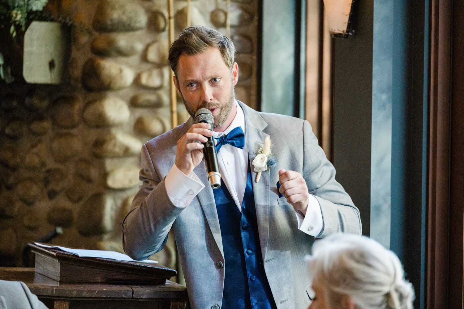 A man in a gray suit delivers a heartfelt speech at a wedding, holding a microphone while gesturing expressively.