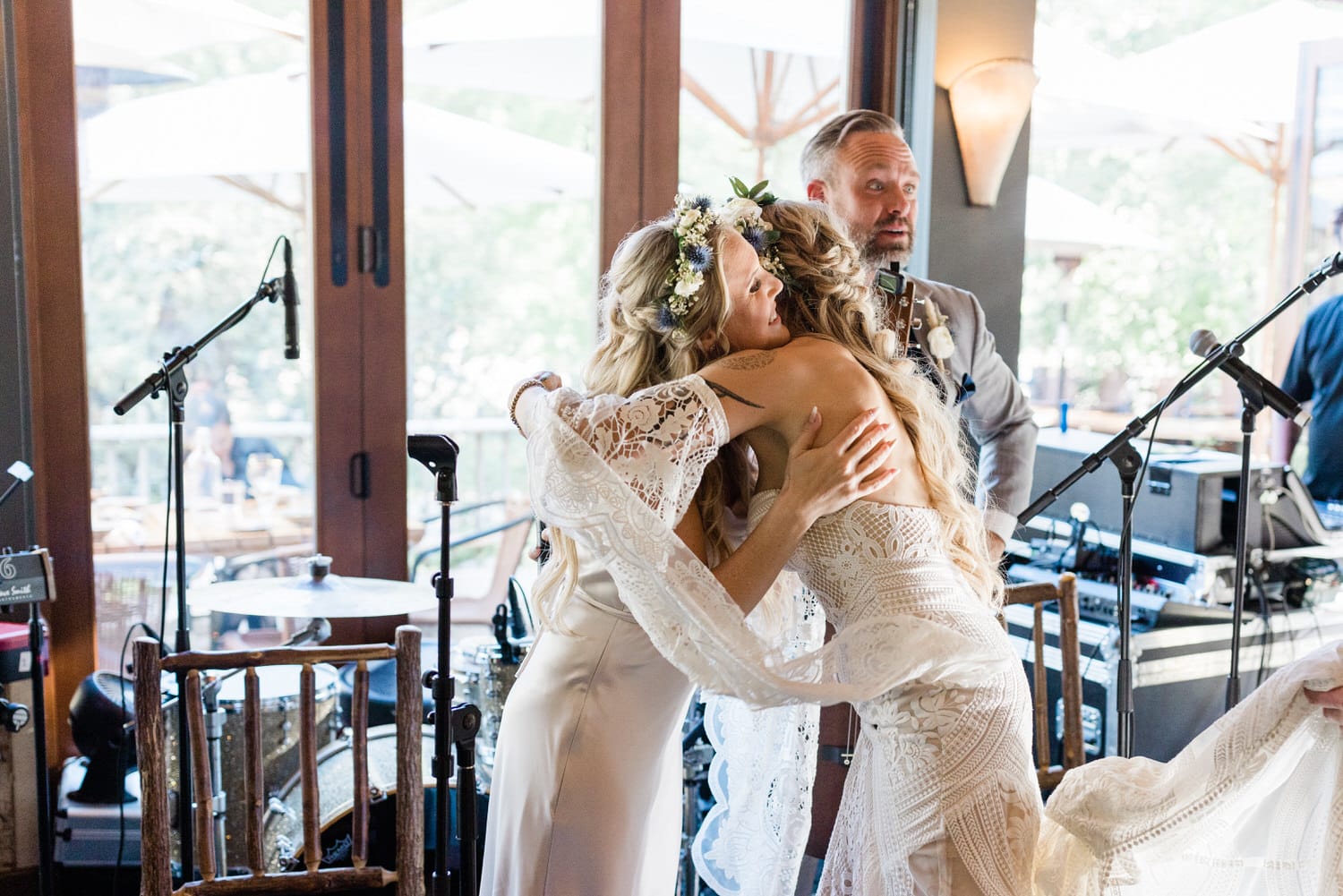 Two brides sharing a heartfelt hug in an intimate wedding setting, with a musician in the background and a lively atmosphere.