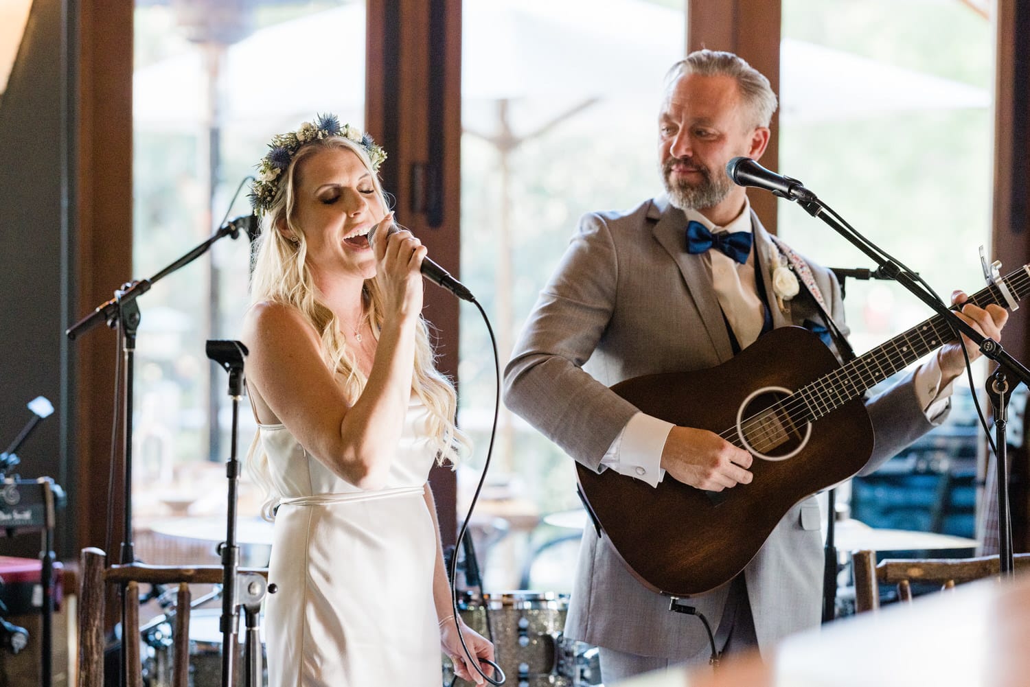 A bride sings passionately into a microphone while her partner accompanies her on an acoustic guitar during a live performance.