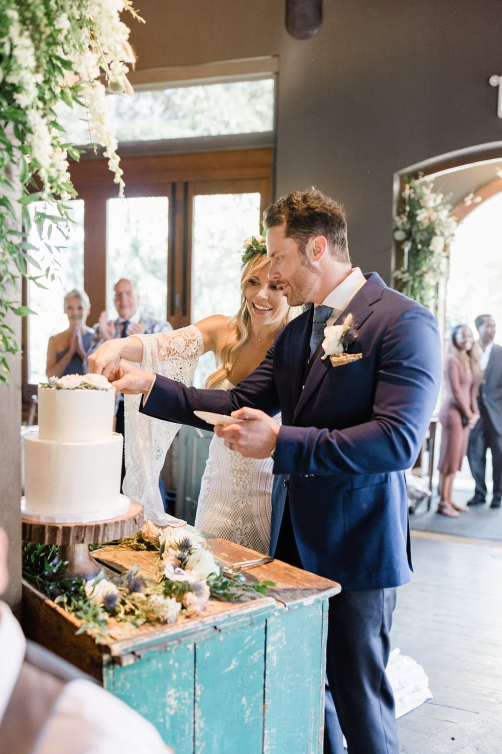 A couple joyfully cutting their wedding cake together, surrounded by a rustic setting and applauding guests.