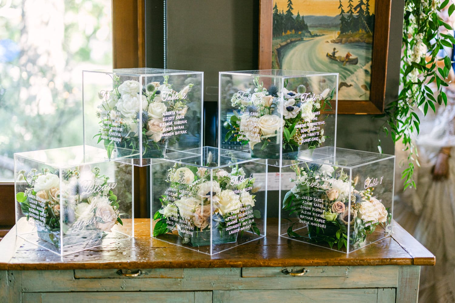 A display of elegant floral arrangements housed in clear acrylic boxes, featuring handwritten table assignments for an event.