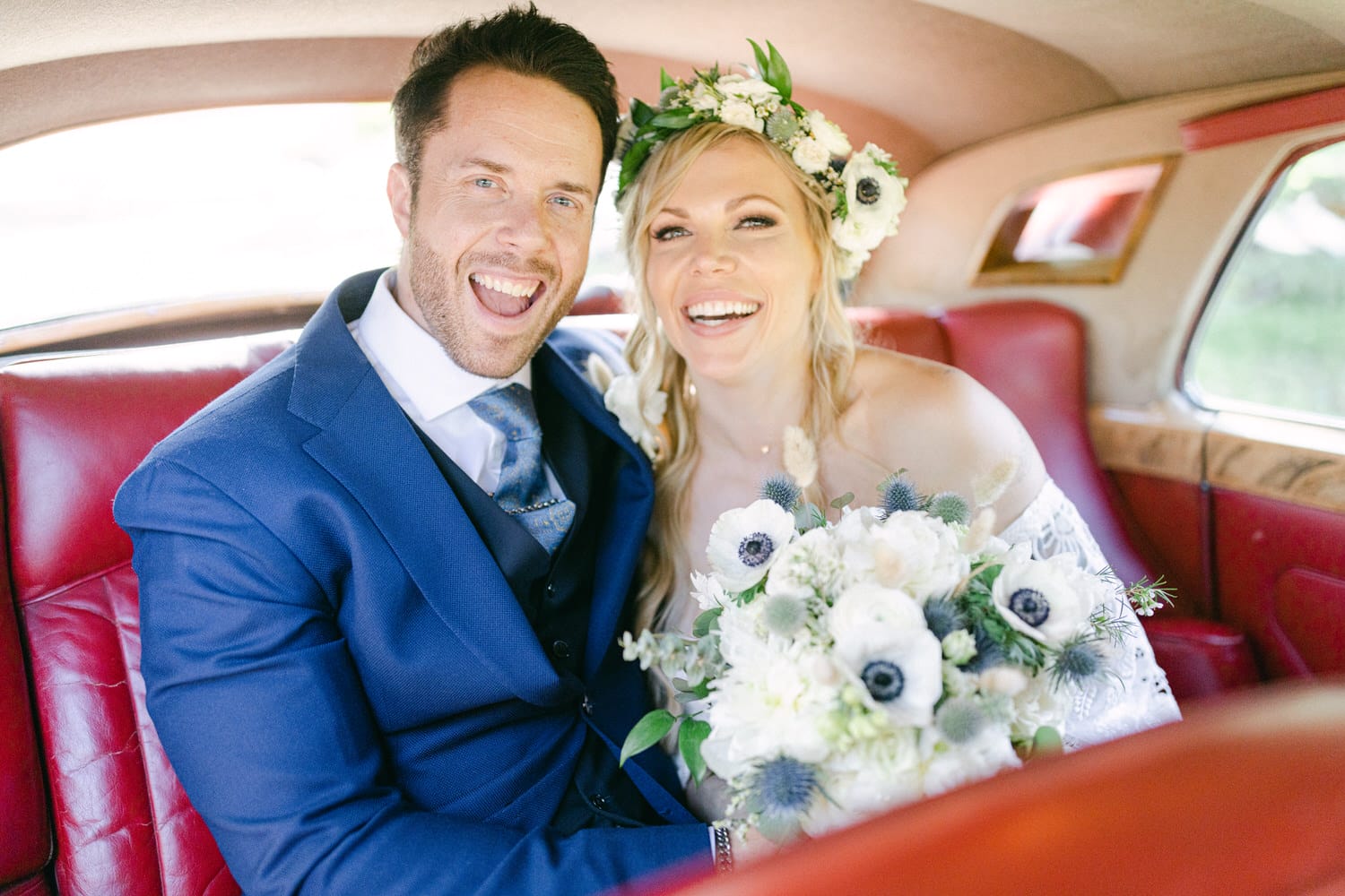 A smiling couple in formal attire sitting in a vintage car, with the bride holding a beautiful bouquet and wearing a floral crown.