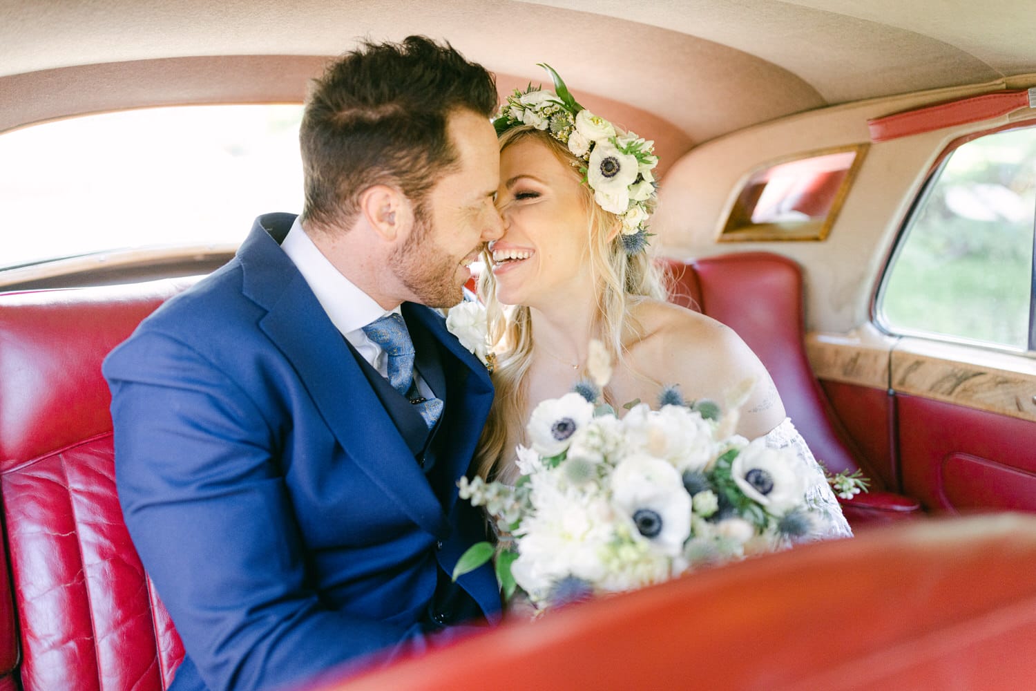 A happy couple sharing a loving smile inside a vintage car, surrounded by beautiful floral arrangements.