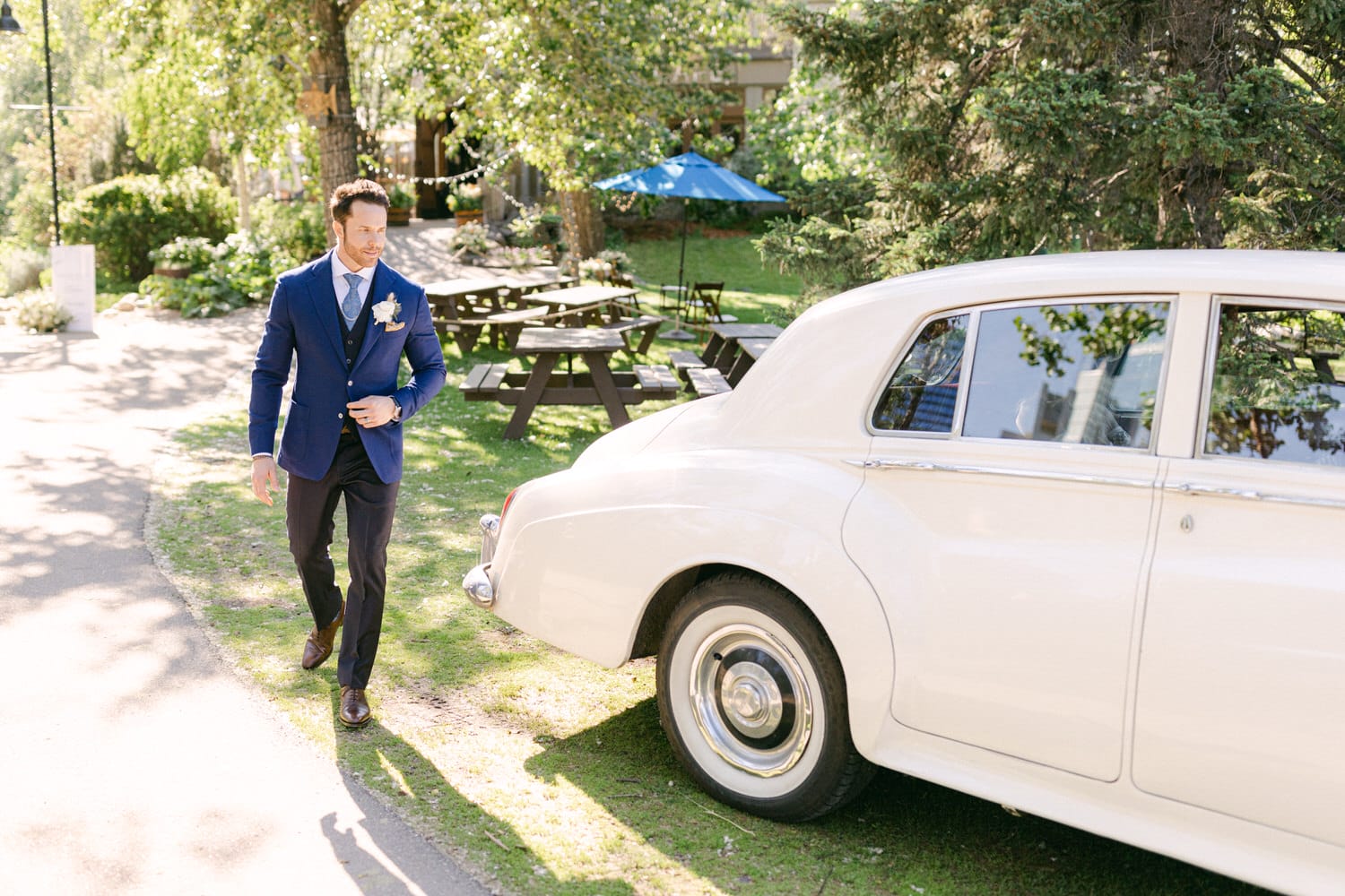 A well-dressed man in a blue suit walks towards a vintage white car in a sunny outdoor setting with trees and picnic tables in the background.