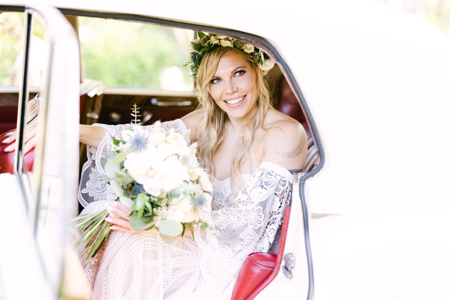 A joyful bride wearing a lace gown and floral crown smiles from the backseat of a classic car, holding a bouquet of flowers.