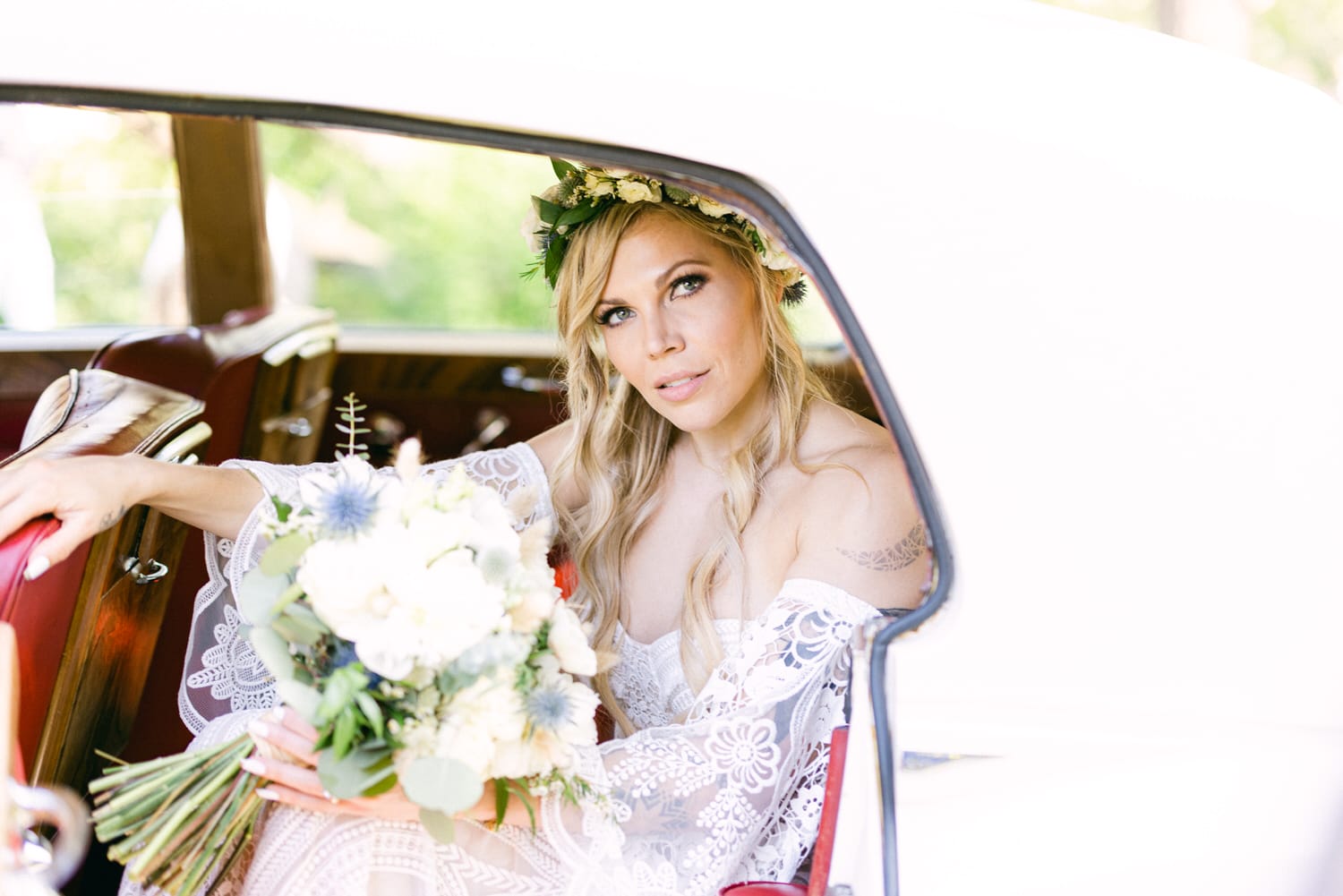 A beautiful bride sitting in a vintage car, holding a bouquet of flowers, adorned with a floral crown, wearing a lace wedding gown.