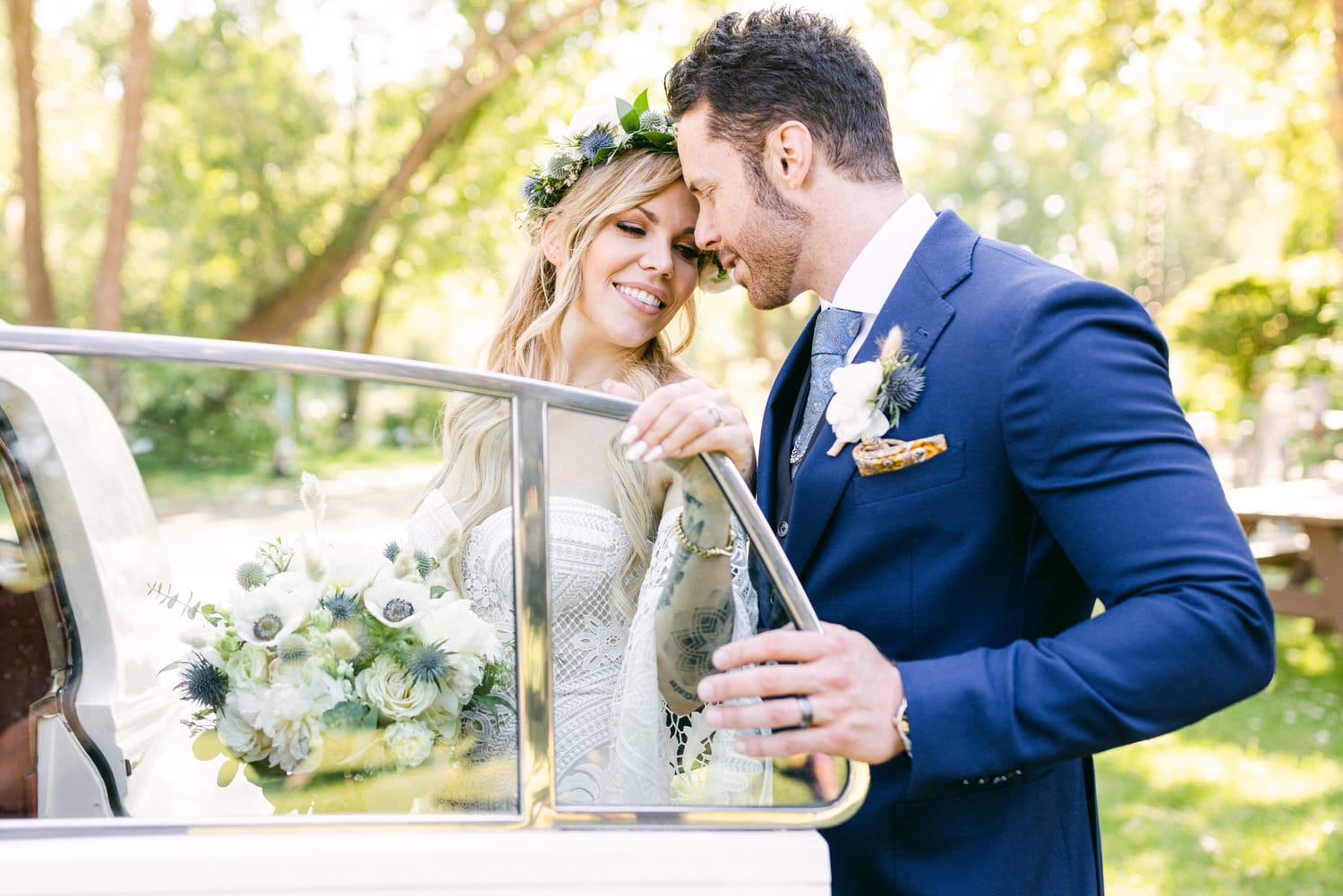 A happy couple sharing a romantic moment by a classic car, surrounded by lush greenery, with the bride holding a beautiful bouquet and wearing a floral crown.