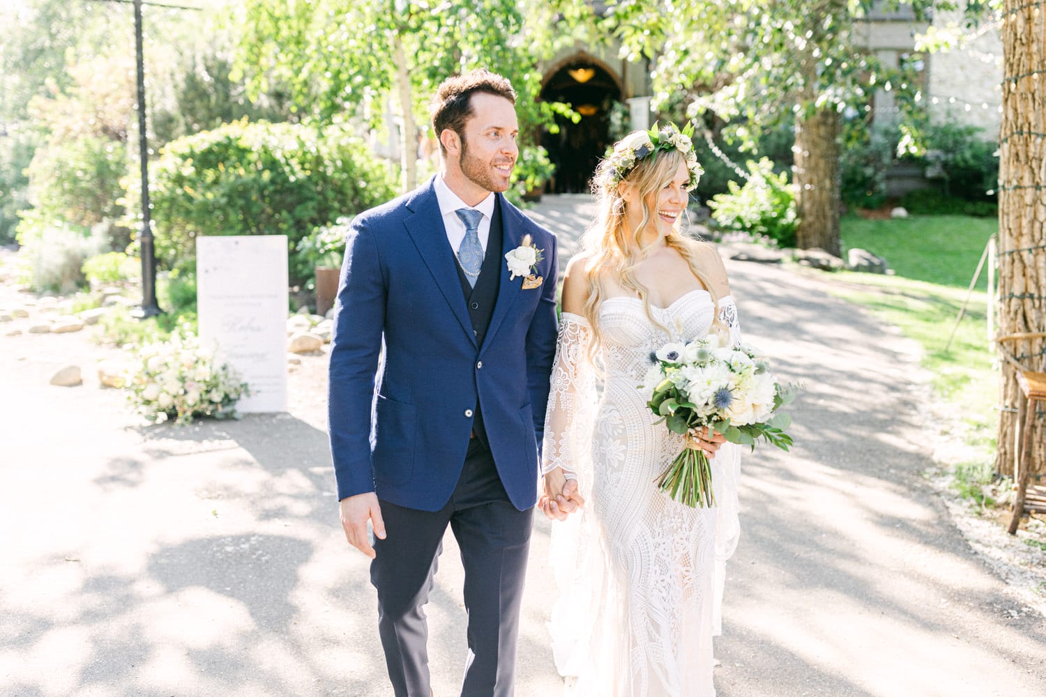 A joyful couple walks hand in hand through a sunlit garden, adorned in elegant wedding attire, surrounded by lush greenery and floral arrangements.