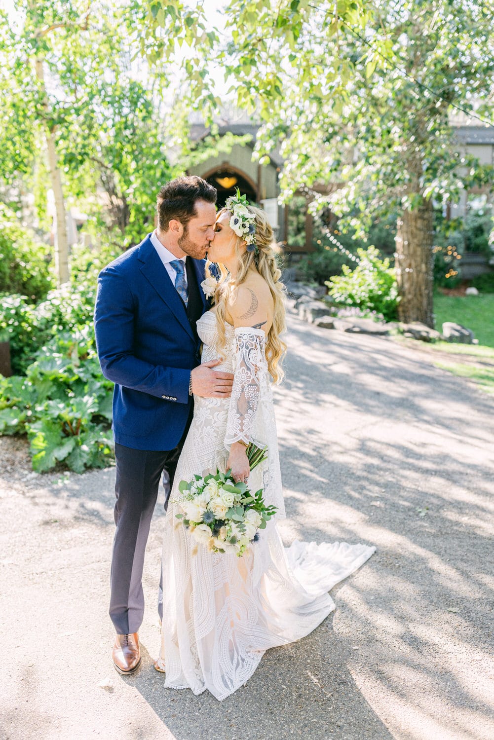 A couple shares a kiss in a lush garden setting, with the bride in a lacy gown and floral crown, and the groom in a blue suit, surrounded by greenery.