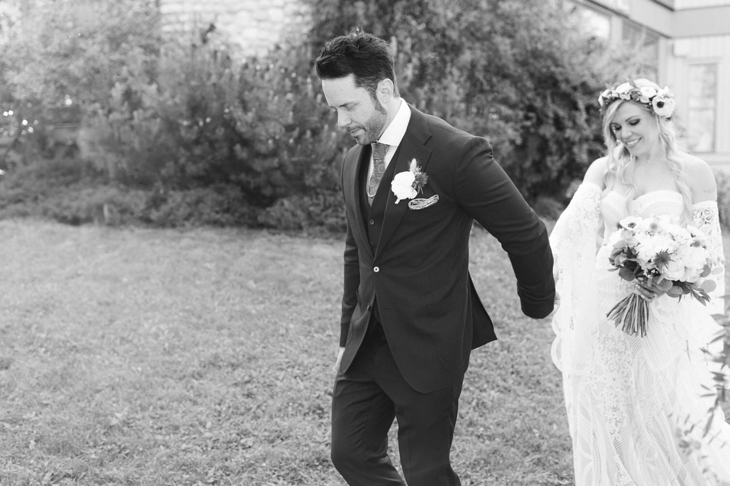 A bride in a lace gown and floral crown holds a bouquet, walking hand-in-hand with a suited groom in a serene outdoor setting.