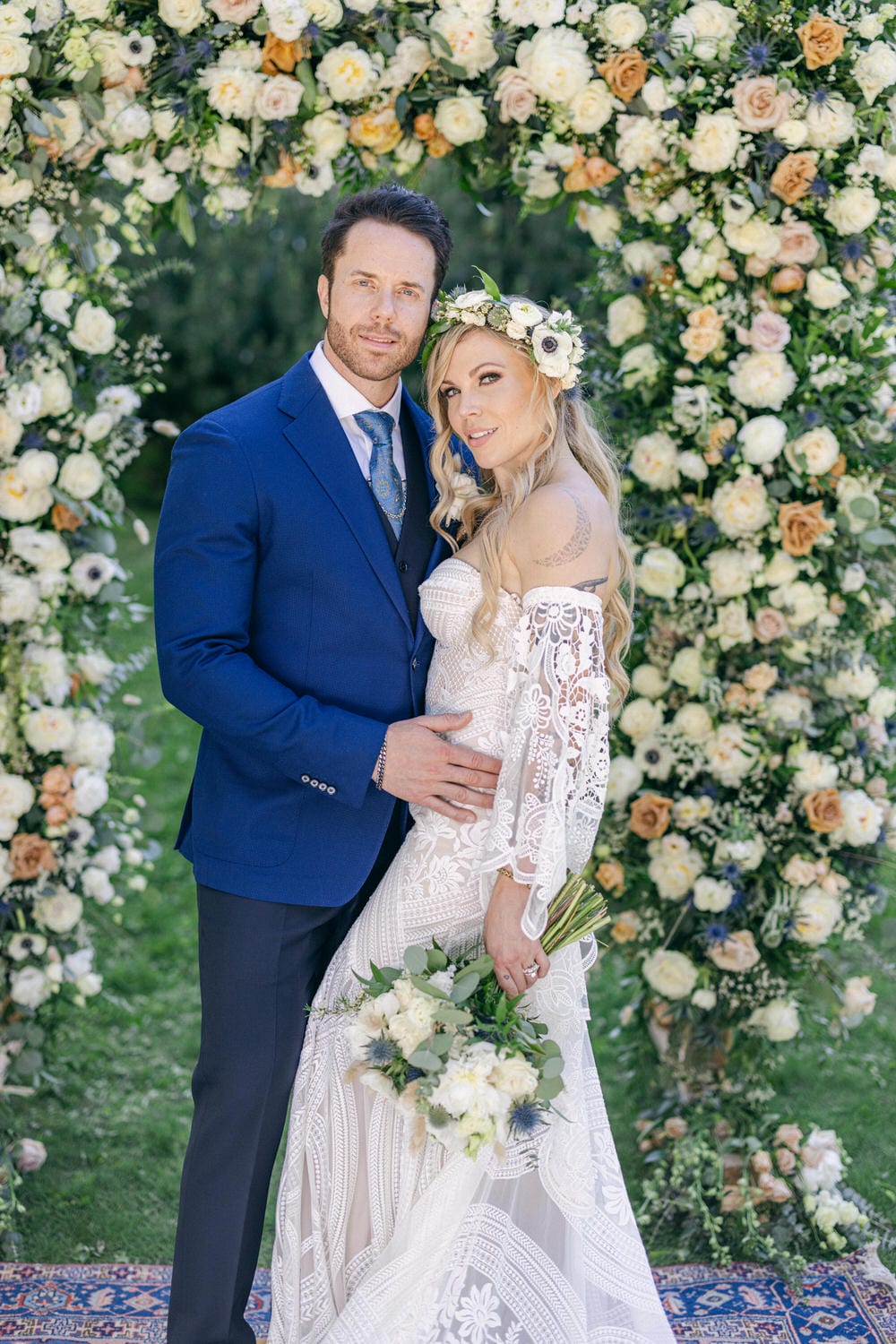 A happy couple poses together in front of a lush floral backdrop, showcasing the bride in a delicate lace gown and floral crown, and the groom in a stylish blue suit.