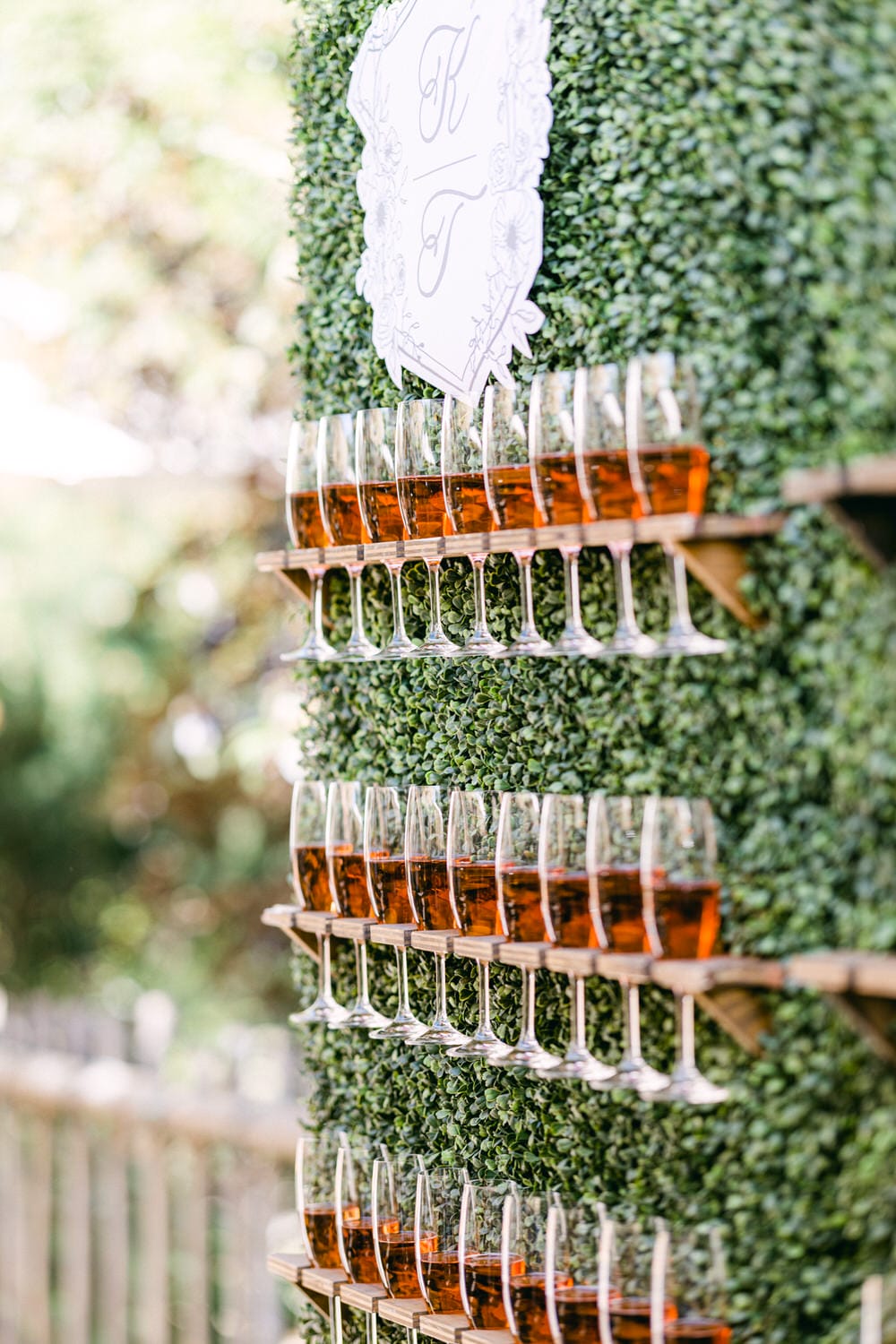 A tiered arrangement of sparkling glasses filled with a reddish-brown drink, set against a lush green backdrop with a decorative sign.