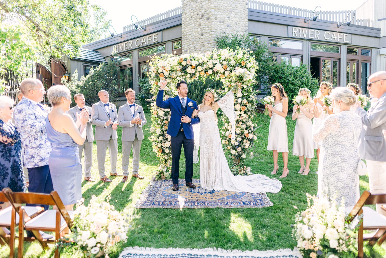 A couple joyfully celebrates their wedding ceremony outdoors, surrounded by guests and a floral archway at the River Cafe.