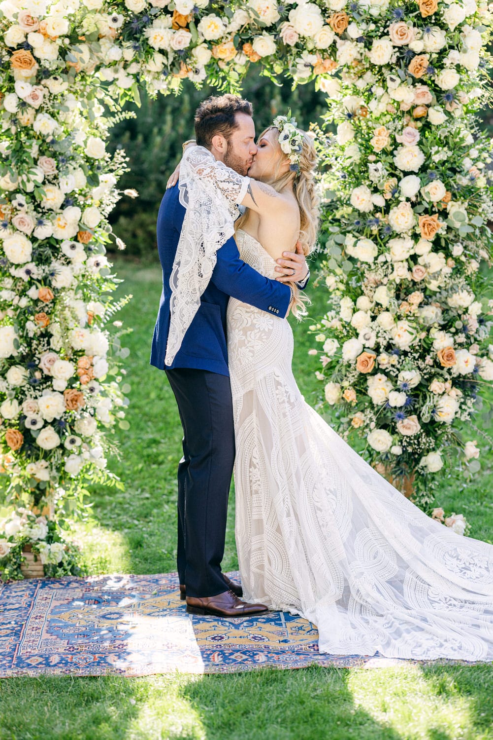 A couple sharing a kiss beneath a floral arch during their wedding ceremony, surrounded by lush greenery and a decorative rug.