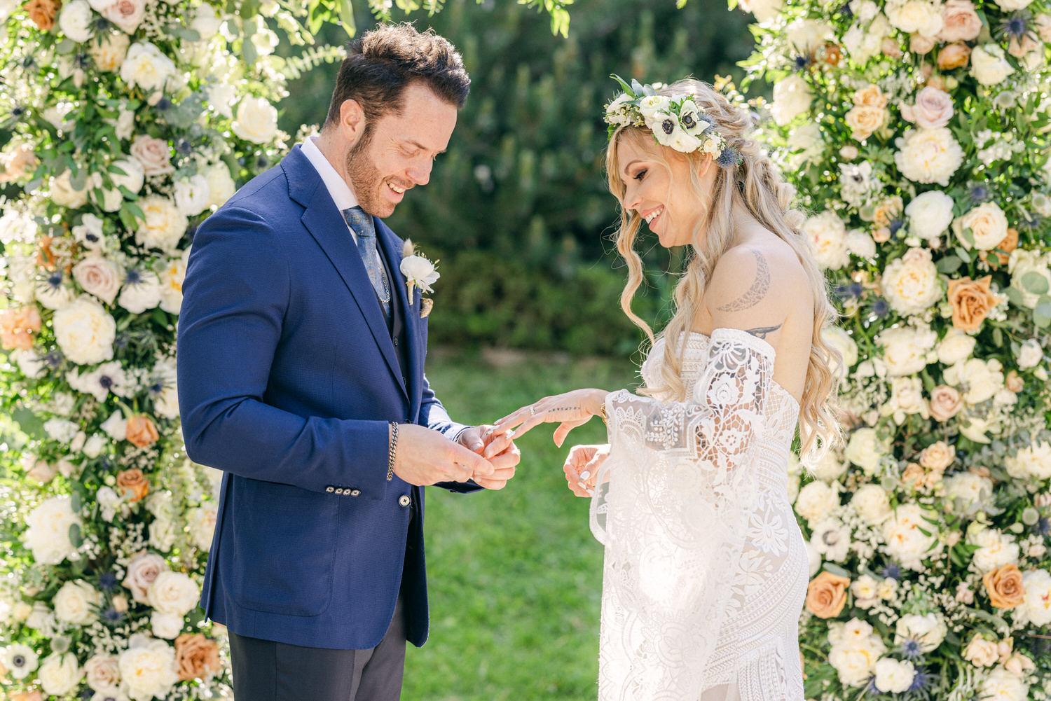 A joyful couple exchanges rings during their outdoor wedding ceremony, surrounded by floral arrangements.