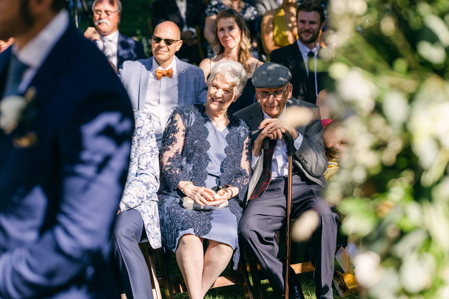 Elderly couple enjoying a wedding ceremony, surrounded by friends and family in a picturesque outdoor setting.