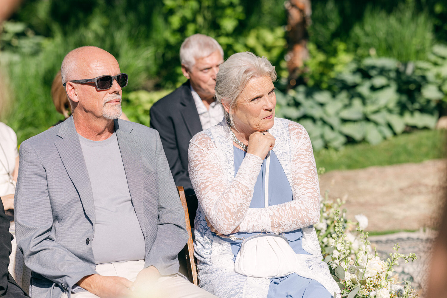 A man in sunglasses and a woman in a lacy white cover-up sit together, attentively observing a wedding ceremony in a lush garden.