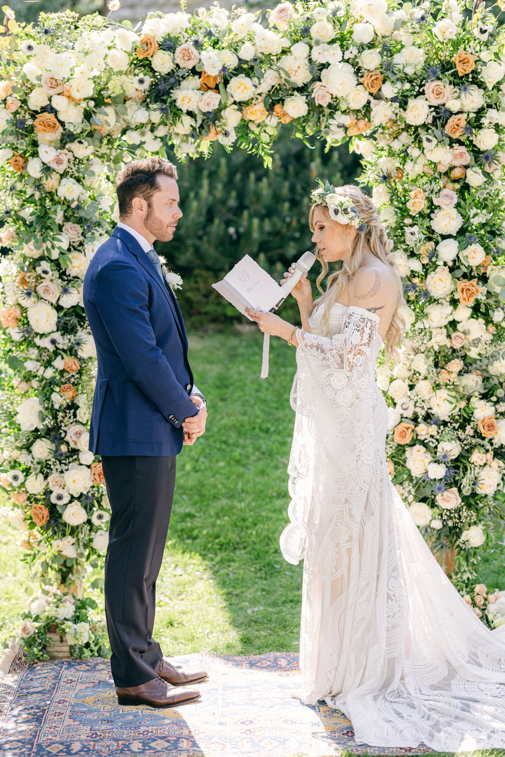 A bride and groom stand before a floral arch during their wedding ceremony, as the bride reads from a booklet while holding a microphone.