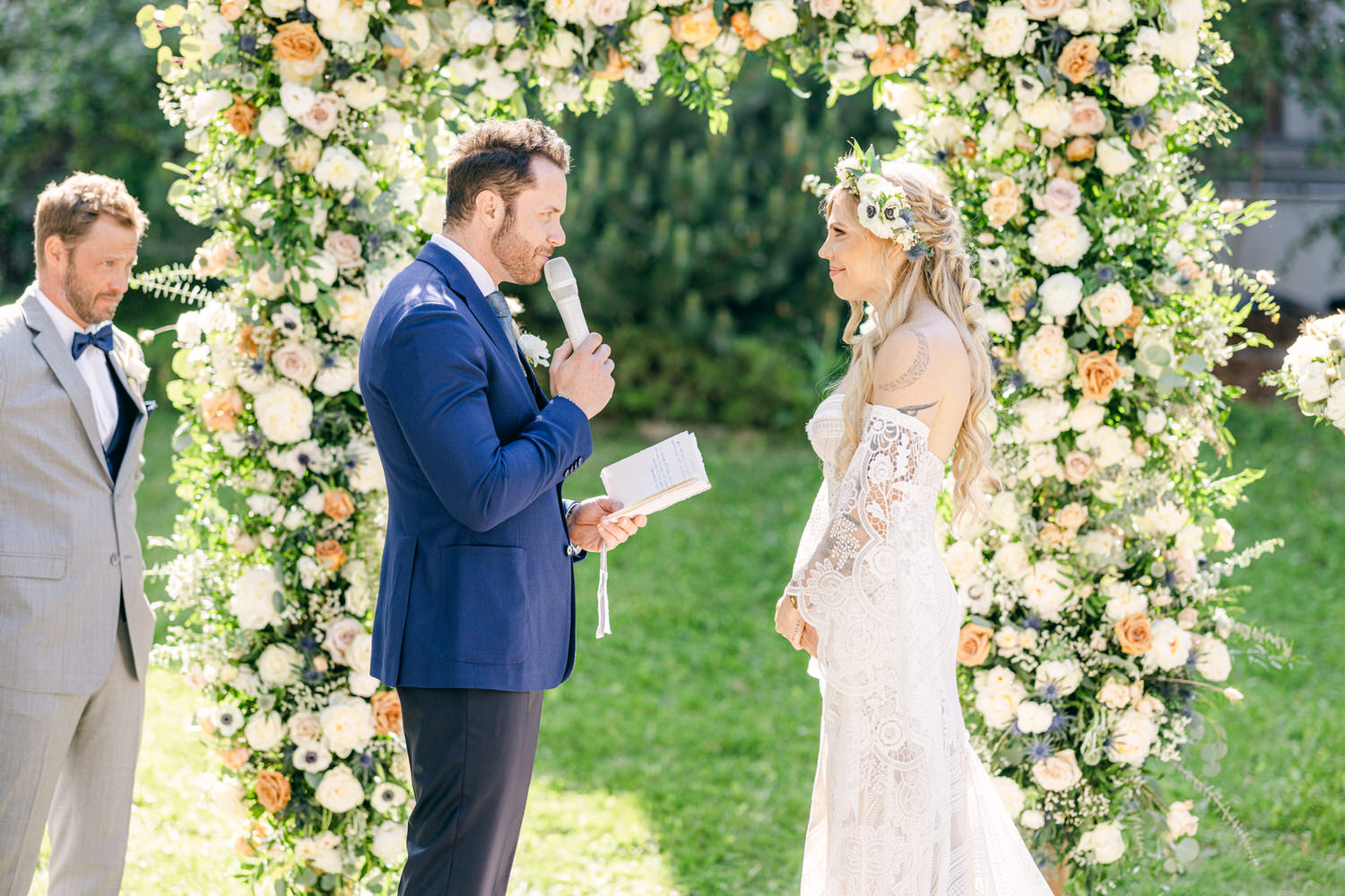 A couple exchanges vows during their wedding ceremony, surrounded by a lush floral arch in a sunny outdoor setting.