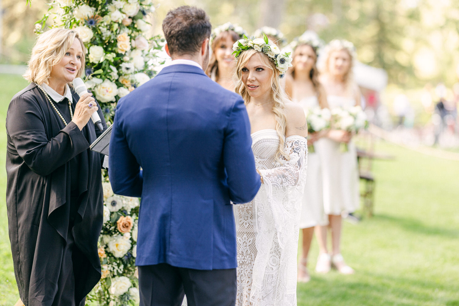 Wedding Ceremony Moments::A bride and groom exchange vows with a celebrant and bridal party in a picturesque outdoor setting, surrounded by floral arrangements.