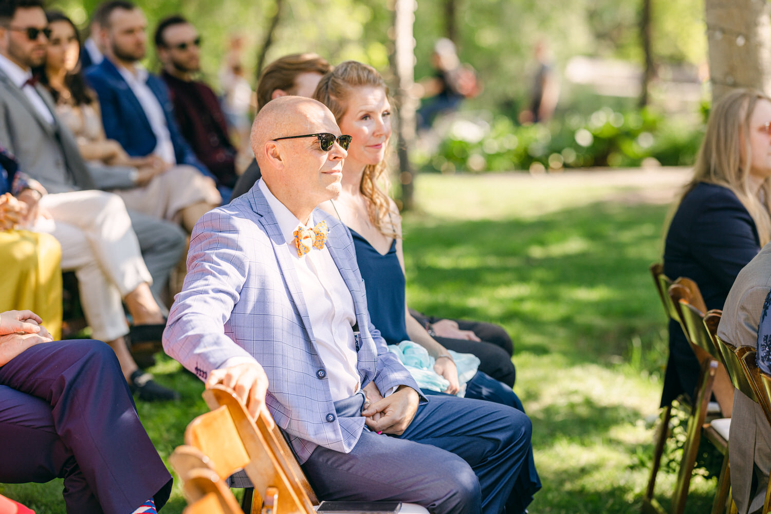 A man in a light blue checkered suit and sunglasses sits among wedding guests in a sunny outdoor setting, with lush greenery in the background.