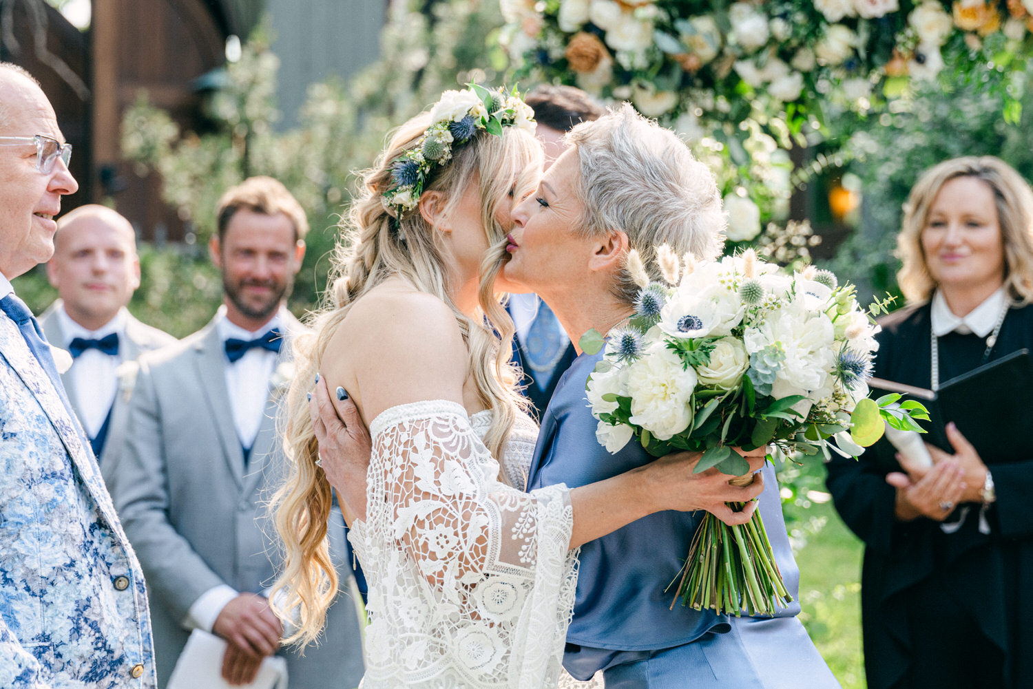 A bride shares a heartfelt kiss with her mother during a wedding ceremony while surrounded by family and friends.