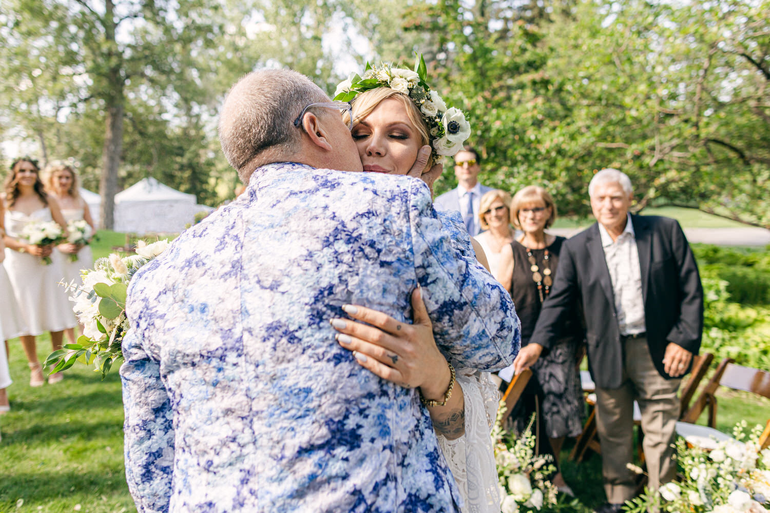 A bride is warmly embraced by her father during an outdoor wedding ceremony, surrounded by guests and natural greenery.