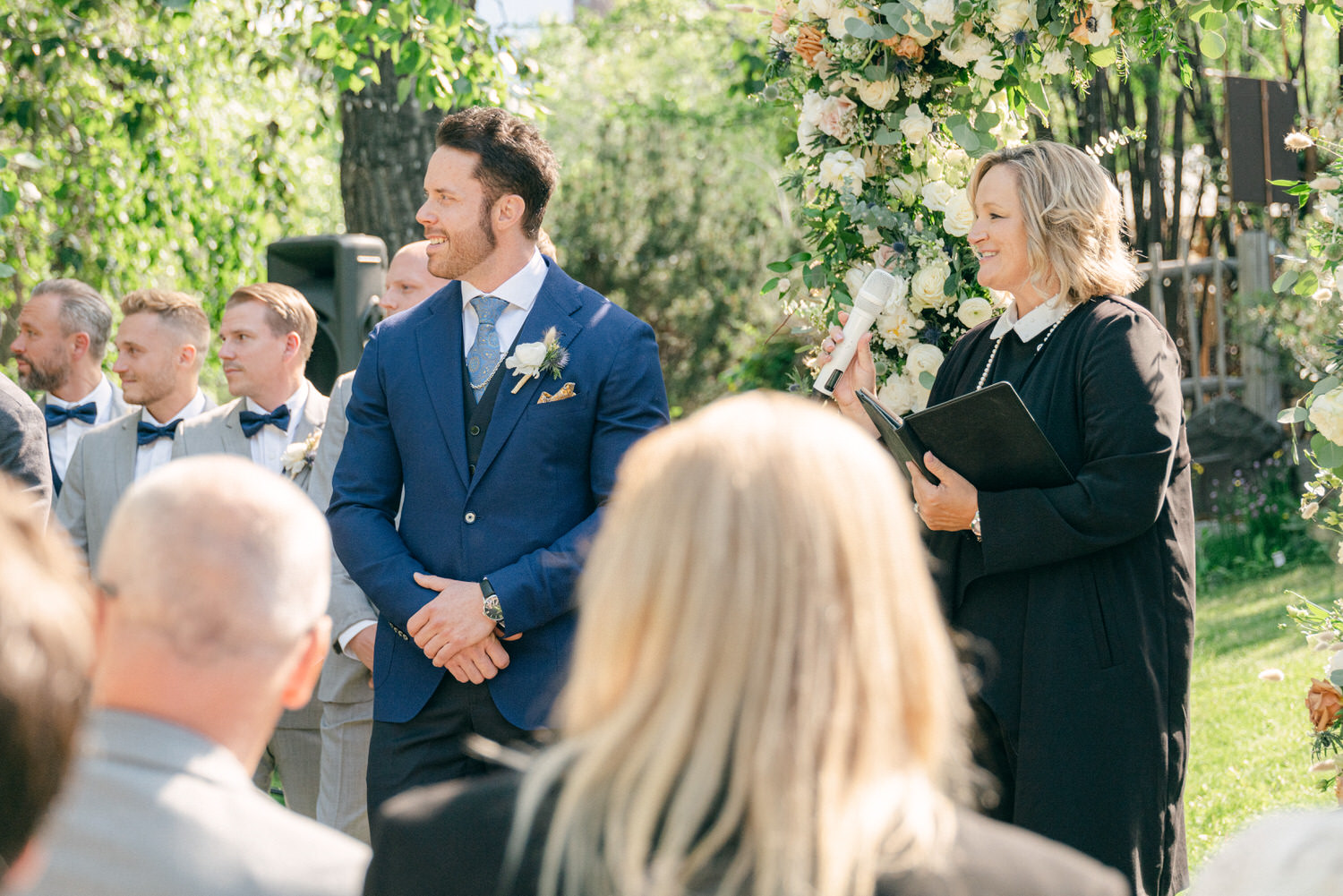 A groom stands attentively as an officiant speaks during a wedding ceremony, surrounded by guests and floral decorations.