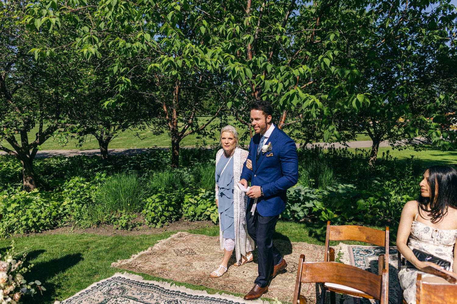 A man in a blue suit escorts a woman in a light blue dress, both smiling, through a lush garden with trees and greenery, as guests look on.